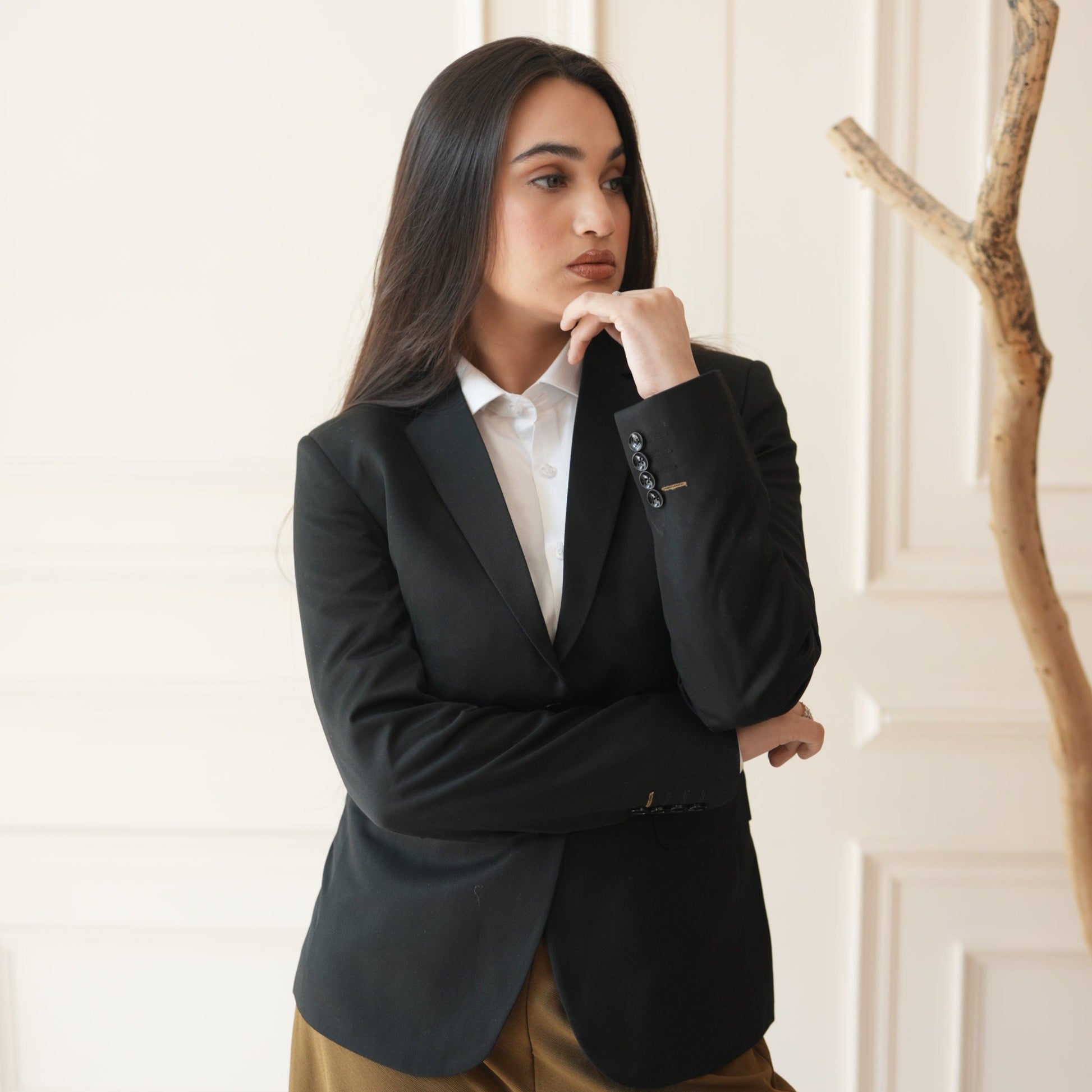 Woman in a black blazer and brown pants standing indoors with a neutral background
