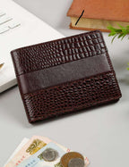 A dark brown bi-fold wallet with a textured crocodile pattern and a smooth horizontal stripe, displayed on a light gray desk with a laptop, book, and coins nearby.