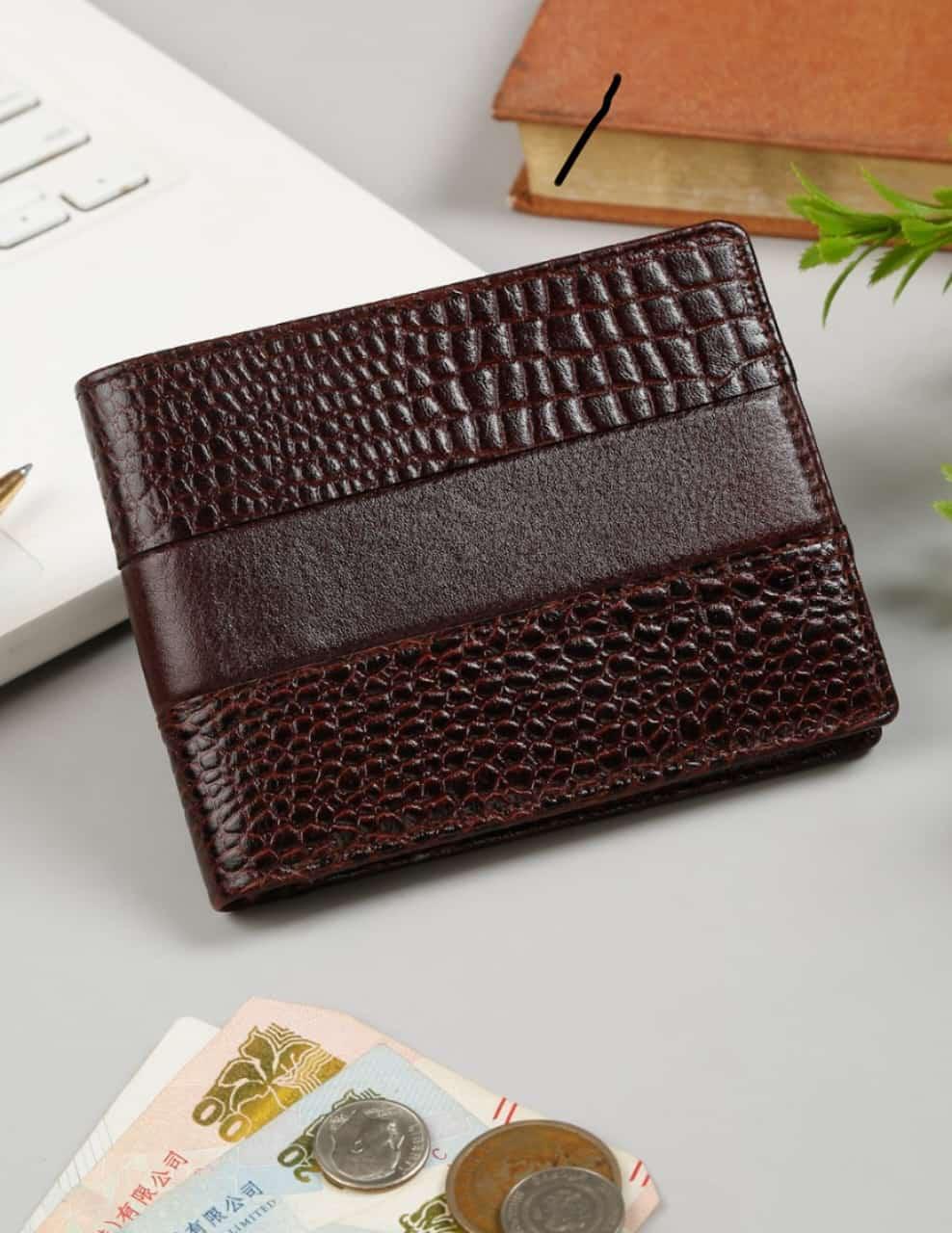 A dark brown bi-fold wallet with a textured crocodile pattern and a smooth horizontal stripe, displayed on a light gray desk with a laptop, book, and coins nearby.